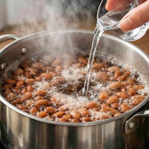 Aquí tienes los textos alternativos (Alt Text) optimizados tanto para accesibilidad como para SEO, centrados en las palabras clave de tus trucos:
1. Para la imagen de las legumbres (Truco nº 2)

    Texto alternativo: "Mano vertiendo un chorro de agua fría en una olla con alubias hirviendo para asustar las legumbres y evitar que se rompan.