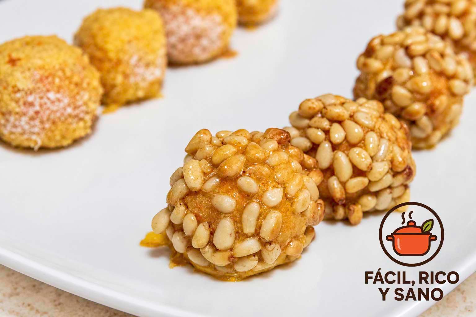 Panellets de boniato rebozados en piñones, con panellets de coco al fondo, sobre un plato blanco