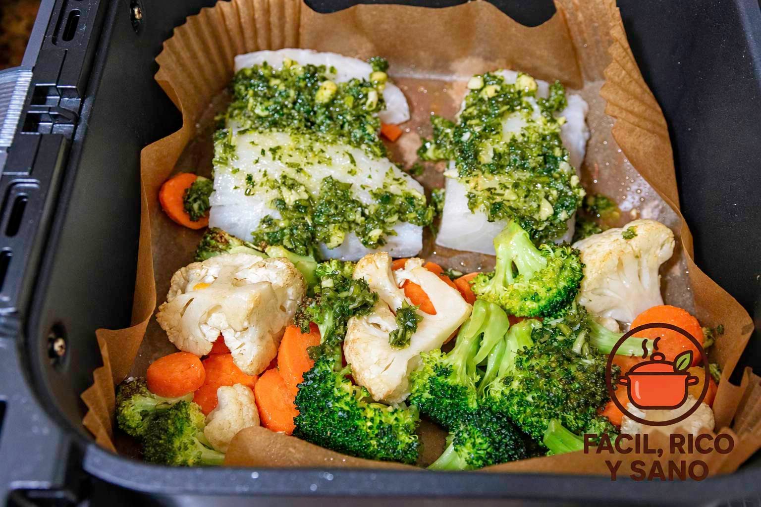 Lomos de bacalao con picada de ajo y perejil junto a brócoli, coliflor y zanahoria en la cesta de la air fryer, sobre papel de horno