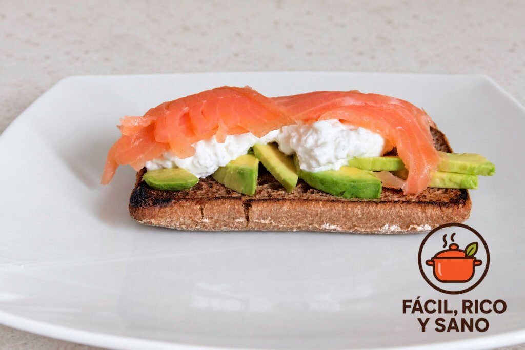 Tostada de aguacate y salmón ahumado con queso cottage sobre pan integral, en un plato blanco